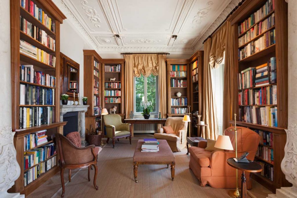 A room lined with full bookshelves, with armchairs, a footstool and a tall curtained window