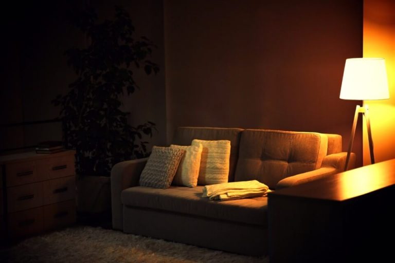 A dim living room with a couch, cushions, a folded blanket and a lit floor lamp