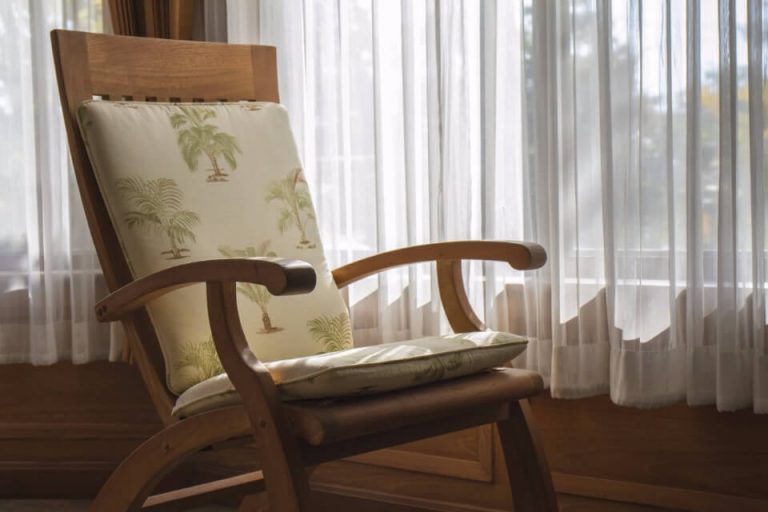 A wooden armchair with a palm-print cushion beside sheer white curtains in soft sunlight