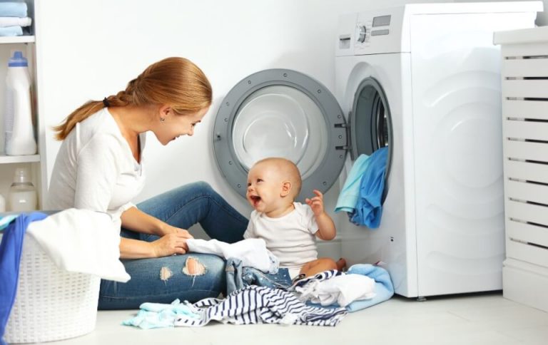 A woman sitting on the floor by a front-loading washing machine with a smiling baby and a pile of laundry