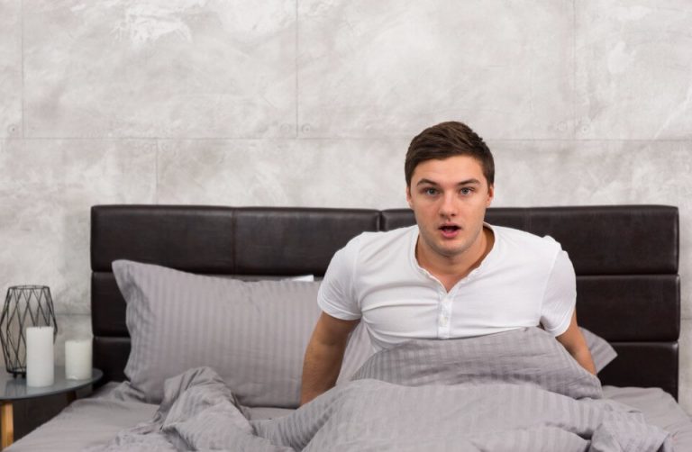 A man sitting up in bed under grey sheets, looking surprised